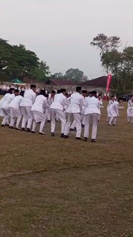 A group of people in white uniforms are performing a coordinated dance routine on a grassy field. They are arranged in a circle and are moving their arms and bodies in unison. Some individuals are holding hands, while others are reaching out towards each other. The background shows trees, buildings, and flags. The sky is overcast.