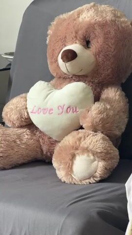 The video displays a close-up shot of a large, plush teddy bear sitting upright on a gray surface, likely a couch or chair. The bear is a light brown color and is holding a white, heart-shaped pillow with pink lettering that reads "Love You." The bear's paws are positioned on either side of the pillow, and its head is tilted slightly to the side. The background is blurred, but it appears to be an indoor setting with a neutral color palette. The lighting is soft and diffused. The video is very short, lasting only a few seconds, and there is no visible movement or sound. The focus is entirely on the teddy bear and the heart-shaped pillow.