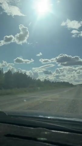 The video shows a view from inside a vehicle, looking forward through the windshield. The road ahead is a dirt or gravel road that curves slightly to the right. The road is bordered by green vegetation and trees on the left, and vegetation and a flat horizon on the right. The sky is blue with scattered white clouds, and the sun is shining brightly, creating lens flare. The interior of the vehicle is visible at the bottom of the frame, showing the dashboard. The lighting suggests it is daytime, likely midday.