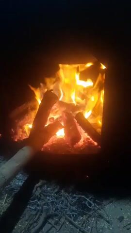 The video shows a close-up of a wood-burning stove at night. The fire inside is bright and active, with flames flickering and logs burning. There is a long stick or branch in the foreground, positioned to the left of the opening of the stove. The ground around the stove is covered in what appears to be dry grass or twigs. The overall scene is dark, with the fire being the primary source of light.