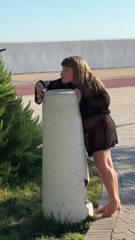 A young girl with long brown hair, wearing a black mesh cover-up and a pink swimsuit, is drinking from a public water fountain. She is standing outdoors on a paved area with a small green bush to her left. Behind her is a white wall and a clear blue sky, suggesting a sunny day. The fountain is made of concrete and is tall and cylindrical. The girl is leaning on the fountain, her head tilted back as she drinks. Her expression is neutral. The scene appears to be a public space, possibly near a beach or park, given the presence of the fountain and the sunny weather. The time of day appears to be late afternoon or early evening, indicated by the warm lighting and long shadows. There is no visible text in the media file.