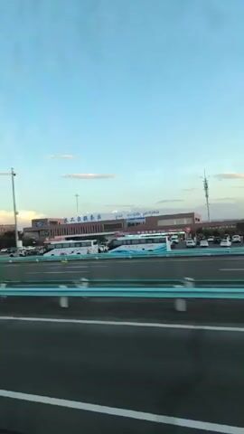 The video shows a view from inside a vehicle, likely a bus or car, looking out at a highway rest stop or service area. The time appears to be late afternoon or early evening, with a clear blue sky and scattered clouds. The rest stop has several buildings, including what appears to be a gas station with pumps and a sign that reads "LNG." There are various vehicles present, including buses, cars, and trucks. The road in the foreground is a highway with a turquoise blue barrier separating lanes. The camera is moving, indicated by the motion blur of passing vehicles and the scenery.