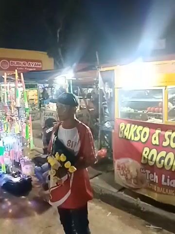 The video shows an outdoor scene at night, likely a market or street fair. In the foreground, a person wearing a red shirt and a black cap stands holding a bunch of yellow objects on sticks. To the left, a display of colorful items on tall stands is illuminated by lights, with some items appearing to be candy or small toys. Behind this display, a stall with the sign "BUBUR AYAM" (Chicken Porridge) is visible. To the right, a food stall with a yellow awning displays a banner that reads "BAKSO & S..." (meatballs and...). Several people are visible in the background, some sitting and others standing near the stalls. The ground appears to be dirt or gravel. The overall atmosphere is lively and commercial.