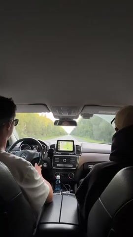 The video shows the interior of a car driving down a road. Two people are inside the car. One person is driving, and the other is in the passenger seat. The driver is wearing a white t-shirt and sunglasses. The passenger is wearing a black hoodie and a baseball cap. The car has a navigation system that shows a map of the road ahead. The road is lined with trees. The sky is overcast. The video is shot from the perspective of someone in the back seat.