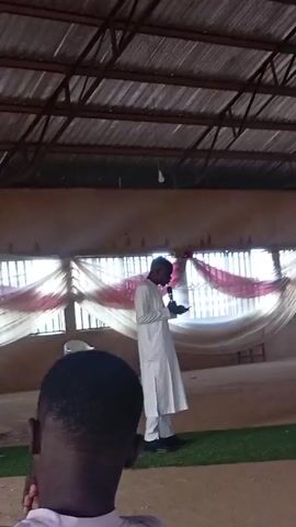 A man in a white robe stands in front of a window with white and red fabric, speaking into a microphone. A person with short dark hair is in the foreground with their back to the camera. The roof is corrugated metal.