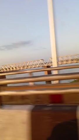 This media file is a video, captured from a moving vehicle, looking out over a wide river. The primary subject is a long, white truss bridge spanning the river. The bridge has multiple sections supported by stone pillars. The water below appears calm, with a slight ripple. A small, dark boat is visible on the river.

In the foreground, there are thick, horizontal railings, likely from the vehicle the camera is in, which are out of focus and tinted in warm, earthy tones. The sky is a pale blue with a few wispy clouds, suggesting it is either early morning or late afternoon. The lighting is soft, indicating a time close to sunrise or sunset. There is no visible text in the media.

The overall scene is peaceful and evokes a sense of travel and transit. The setting appears to be a natural landscape with a significant man-made structure, the bridge, dominating the view across the water. The motion blur from the moving vehicle adds a dynamic element to the static scenery. Based on the provided location context, the scene is Jebba, Nigeria, likely depicting the Jebba Bridge over the River Niger.
