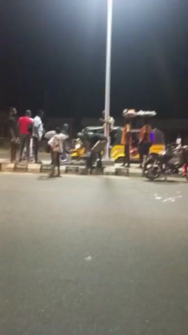 At night, under the illumination of a street light, a group of about a dozen people are gathered on a sidewalk and in the street, seemingly attending to a yellow auto rickshaw (tuk-tuk). Some people are close to the tuk-tuk, while others are standing further back. In the foreground, a dark motorcycle lies on its side on the sidewalk. A car passes by in the street. The environment appears to be an urban or semi-urban setting with buildings visible in the background. The overall scene suggests the aftermath of an incident, possibly a minor accident or breakdown involving the tuk-tuk.