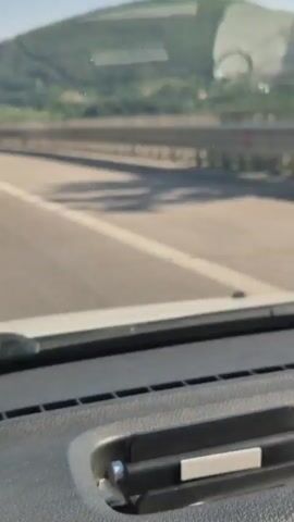 A first-person perspective video shows a view from inside a vehicle driving on a highway. The road ahead is visible, with a white line marking the lane. To the right of the highway, there is a guardrail and a sloping green hill. The sky is clear and blue. In the distance, the hill is covered with trees, and there are some structures visible on the hillside. The vehicle appears to be moving at a moderate speed. The video is short, lasting only a few seconds.