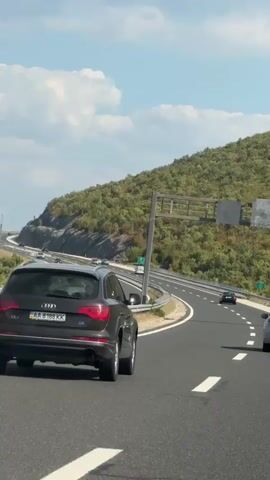 A dark colored Audi SUV is traveling on a highway. The SUV is in the right lane. A white compact car is in the left lane. The highway is surrounded by green, tree-covered hills. The sky is blue with some white clouds. There are other cars on the highway. The time is daytime.