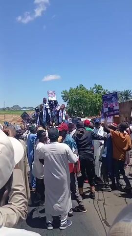 A group of people are gathered outdoors on a road under a clear blue sky. Many of them are holding up posters with images of people on them, likely for a political campaign. Some individuals are wearing traditional clothing, while others are in more casual attire. One person in the center appears to be wearing black tactical gear and holding a baton. The overall atmosphere suggests a rally or protest. The ground is paved, and there are trees and some buildings visible in the background.