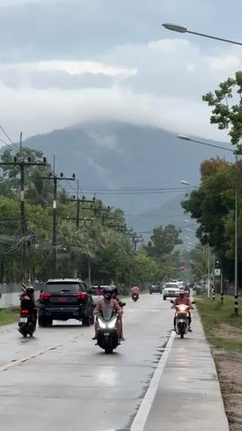 The video shows a road in Thailand on a cloudy day, with a mountain covered in mist in the background.  The road is wet, likely from a recent rain. Traffic moves in both directions, including cars and scooters.  A woman wearing a pink shirt and cap is riding a white scooter towards the camera.  Power lines stretch across the road overhead.  Trees and vegetation line the sides of the road.