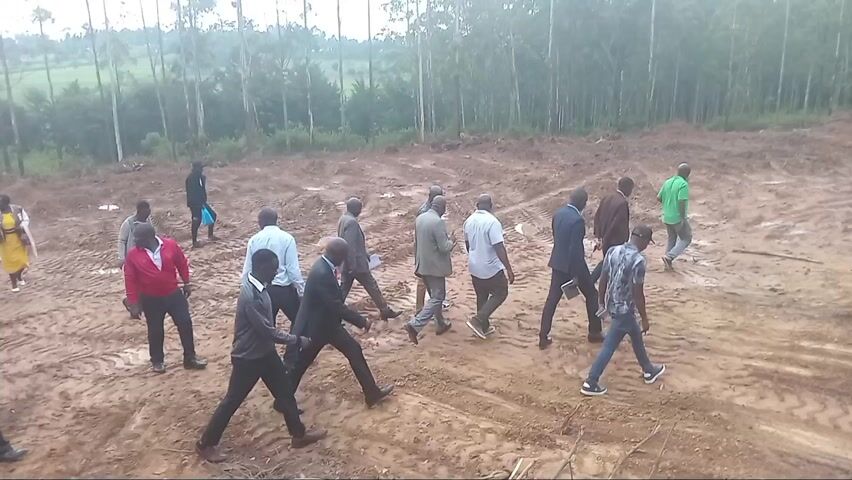 The video captures a group of approximately 15-20 individuals traversing a large expanse of recently cleared, muddy land. The reddish-brown soil is heavily churned with distinct tire tracks, indicating recent activity by heavy machinery. Scattered logs and branches are visible on the uneven surface.

In the background, a dense stand of tall, slender trees, consistent with eucalyptus species, forms a prominent tree line. Beyond these trees, hints of green fields and rolling hills are visible under an overcast sky, suggesting a cloudy day.

The group is predominantly composed of men, with a few women interspersed, all walking across the terrain, generally moving from left to right across the frame. Their attire varies; some wear formal suits or blazers, while others are in more casual shirts and trousers. One woman is seen in a bright yellow dress, another individual in a red shirt, and one man wears a reflective safety vest.

The individuals appear to be conducting an inspection or site visit of the cleared area, looking around and engaging in conversation as they walk. The overall atmosphere suggests a purposeful and collaborative assessment of the land. Given the context of Moticho ward, Kenya, this scene likely relates to a land development project, infrastructure assessment, or agricultural initiative.

No discernible text is visible within the frame.