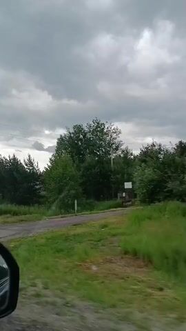 The video is a first-person perspective of a drive down a paved road under a cloudy sky. The road is bordered by tall green grass and trees. A sign with Cyrillic lettering is visible on the left side of the road, followed by a crosswalk. In the distance, houses and a bus stop can be seen. The drive appears to be in a rural or semi-rural setting.