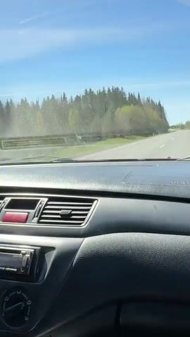 The view from inside a car driving on a paved road with green trees and a clear blue sky in the background. The car's dashboard is visible, including the air vents. The windshield is slightly dirty. The road appears to be relatively empty.