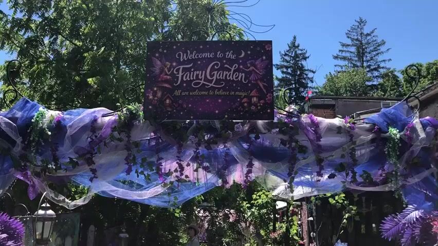 The video shows an outdoor scene with a sign that says "Welcome to the Fairy Garden. All are welcome to believe in magic!" The sign is decorated with purple and pink colors and features images of fairies and mushroom houses. The entrance to the fairy garden is decorated with flowing purple and blue fabrics and artificial greenery. In the background, there is a white building and trees. There are also lanterns and other decorations that suggest a whimsical and magical atmosphere. A person can be seen in the background, walking through the fairy garden.