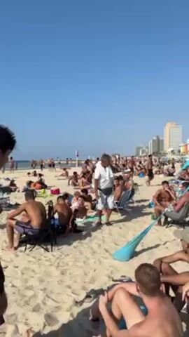 The video shows a crowded beach with people sunbathing and relaxing. The sky is clear and blue. A man in a white shirt and light-colored shorts walks through the crowd, carrying a cooler bag with the words "The 6 best!!!" written on it in Hebrew. He appears to be selling drinks. The man has a fanny pack around his waist and a towel over his shoulder. In the background, there are tall buildings.