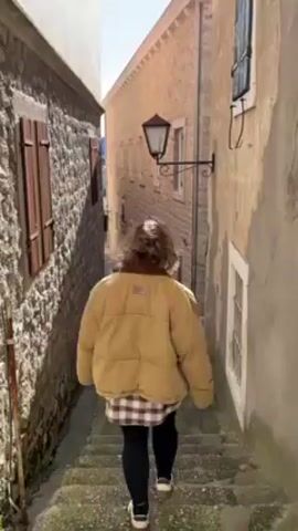 A person is descending a narrow, stone staircase in an alleyway. The person is wearing a tan puffer jacket, a patterned shirt, black leggings, and white sneakers.  The alley is bordered by old stone buildings with windows and shutters. A street lamp is mounted on the wall of one of the buildings. The day appears sunny. The camera follows the person from behind.