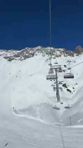 The video shows a view from a ski lift going up a mountain on a sunny day. The sky is clear and blue. In the foreground is the ski lift infrastructure, including the cables and chairs. Below, the ski slope is covered in snow, with tracks from skiers and snowboarders visible.  Several people are skiing and snowboarding down the slope. Red poles mark the edges of the ski run. The background features a snow-covered mountain range.