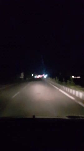 The video shows a night-time drive on a road. The camera is positioned inside a vehicle, looking forward. The road is paved and has white markings. On the left side of the road, there are some trees and vegetation, and on the right, there appears to be a curb or roadside structure. Distant lights from other vehicles are visible, suggesting traffic. The overall scene is dark, indicating it is nighttime. The air appears slightly misty or foggy, as evidenced by the diffuse glow of the headlights. The location is identified as Pudupattanam, India. No people or specific objects are clearly discernible due to the low light conditions and the motion of the vehicle. The activity is a drive through the area at night.