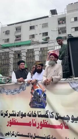 The video shows a group of men standing behind a banner with chains wrapped around a logo that appears to be for "Geo News." The men are dressed in traditional religious attire, including turbans and robes. One man is speaking into a microphone. The banner has text in Urdu script. The background shows a building with balconies and air conditioning units. The sky is overcast. The overall scene appears to be a protest or demonstration.