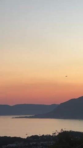This is a vertical video of a sunset over a body of water with mountains in the background. A small airplane is visible flying in the sky. The sky is a gradient of colors from light yellow at the top to orange and pink near the horizon. The water reflects the colors of the sky. Silhouetted mountains line the distant shore, and a small town is visible on the lower left. The overall impression is of a peaceful and beautiful natural scene.