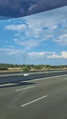 The video shows a view from a moving vehicle on a highway. The sky is blue with scattered white clouds. On the right side of the highway, there is a barrier separating the road from a grassy area with some trees and bushes. In the distance, there are power lines supported by a large tower. Several cars and trucks are driving on the highway in the same direction as the camera. One truck, a red semi-truck with a white trailer, is visible in the foreground on the right lane. The road has white lane markings. The video appears to be filmed during the daytime, likely in the afternoon, given the lighting and cloud formation.