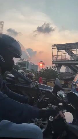 The video shows a street scene at sunset. There is a lot of traffic, with motorcycles and cars. The sky is orange and pink, with some clouds. In the background, there is a pedestrian bridge and some buildings. A person is riding a motorcycle in the foreground, wearing a black helmet and a blue jacket. Another person is riding on the back of the motorcycle. The video is shaky, suggesting it was filmed from a moving vehicle.