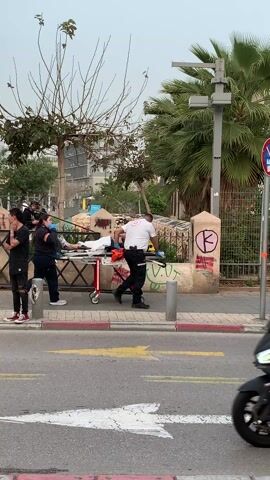 The video shows the scene of an apparent emergency, with paramedics attending to someone on a stretcher next to a yellow ambulance. The paramedics are dressed in emergency medical service uniforms. The person on the stretcher appears to be receiving medical attention. The ambulance has Hebrew writing and the Star of David emblem on it, identifying it as an Israeli ambulance. Various passersby are present, including people on motorcycles and bicycles. The background includes buildings, a palm tree, and a fence with graffiti. It seems to be an urban setting in Israel.