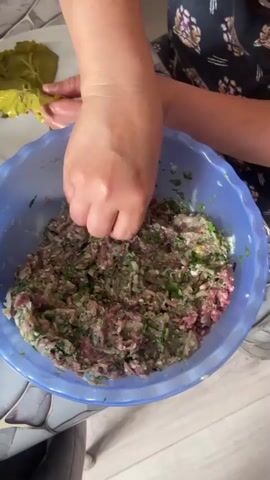 A person is shown in the process of preparing dolmades. Their hands are visible, carefully placing a filling mixture onto a grape leaf, which is held on a white plate. The filling mixture, a combination of ground meat, rice, and herbs, is visible in a large blue bowl in the foreground. The background shows a tiled surface and the person is wearing a patterned shirt. The overall scene depicts a home kitchen environment where food preparation is taking place.