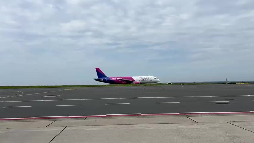 An airplane with a purple and white livery sits on an airport tarmac. The airplane is facing to the right of the frame. The sky is cloudy. The tarmac is gray with white lines painted on it. The camera is stationary.