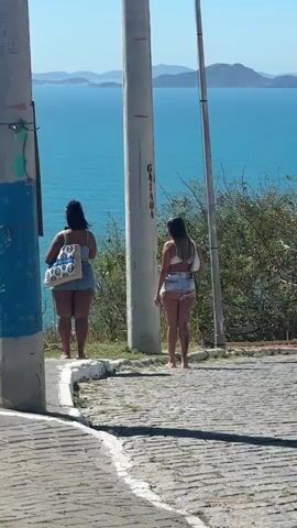 Two women stand facing away from the camera on a paved surface. The woman on the left wears a blue top and denim shorts, carrying a tote bag over her left shoulder. The woman on the right wears a white bikini top and distressed denim shorts. Both women have their backs to the viewer. They are positioned near two utility poles, with a backdrop of a blue ocean and distant hills under a clear sky. The ground is a cobblestone pathway.