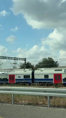 From a moving vehicle, a train is seen traveling along tracks parallel to the road. The train is white with blue stripes and red doors. Overhead electrical lines supply power to the train. The sky is blue with scattered white clouds. A metal guardrail separates the road from the railway. A few sparse plants are visible between the road and the tracks.