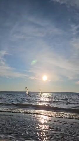 Two people are windsurfing on the ocean. The sun is shining brightly, casting a glare on the water. The sky is blue with some clouds. There are waves on the water. The camera is positioned on the beach, looking out at the ocean.