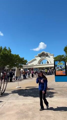 The video shows the entrance to Parque Warner Madrid, with a long line of people waiting to enter. A woman is walking in front of the entrance, talking on her phone. The weather is sunny with a blue sky and a few clouds. There are trees and a large sign for the park. In the background, there are rides and attractions. The video zooms in on the Parque Warner Madrid sign. The camera pans around to show more of the park and the people in line.
