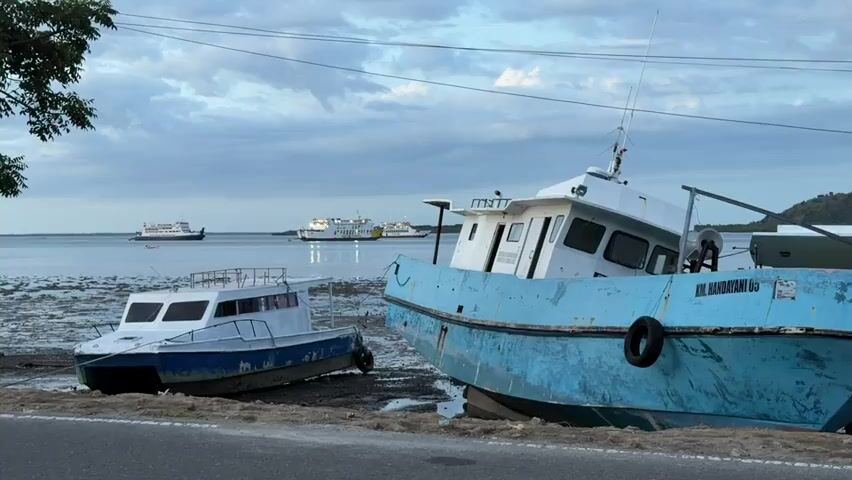 The scene is a coastal area, likely a port or harbor, with a road running alongside the water. Two boats are beached or stranded on the muddy shore near the road. One boat is light blue and white, catamaran style, while the other is a larger, light blue fishing vessel. The name "KM. HANDAYANI 05" is visible on the side of the larger boat. In the distance, several larger ferry or cruise ships are visible on the water. The sky is overcast, suggesting either early morning or late afternoon. A person on a motorcycle drives by on the road in front of the boats. Overhead, there are power lines. A tree branch with leaves is visible on the left side of the frame.