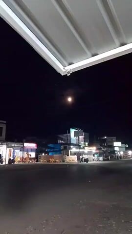 The video shows a nighttime scene with a full moon visible in the dark sky. In the foreground, a portion of a white, corrugated metal awning is visible. Below the awning, a street or road stretches out, with shops and buildings lining the opposite side. The buildings are lit with various colorful lights, suggesting commercial establishments. Several motorcycles are visible on the road, some in motion, indicated by their headlights. There are also some tables and chairs set up outside one of the shops. The overall impression is of a street scene at night, possibly in a town or market area.