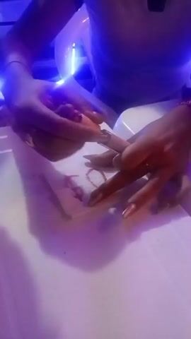 The video captures a close-up view of a person's hands writing on a white paper napkin. The right hand uses a tube of red liquid, likely lipstick or lip gloss, to draw the characters "$20" onto the napkin, while the left hand steadies the paper and briefly holds a small brush. The hands have long, manicured nails, some adorned with light-colored polish. The activity occurs on a white table in what appears to be an indoor setting. The scene is dimly lit with prominent blue and purple neon or LED lighting, suggesting a nighttime environment such as a bar, club, or casual eatery. Part of a person's torso, wearing a light-colored top, is visible in the background. The overall atmosphere appears casual. This scene takes place in Juan Griego, Venezuela.