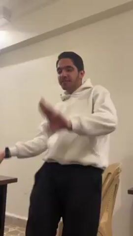 A young man is sitting on a chair in a room, moving his body to the music. He is wearing a white hoodie and black sweatpants. The room has beige walls, a tiled floor, and two small tables. There is a television on a stand in the background. The man is smiling and seems to be enjoying the music.