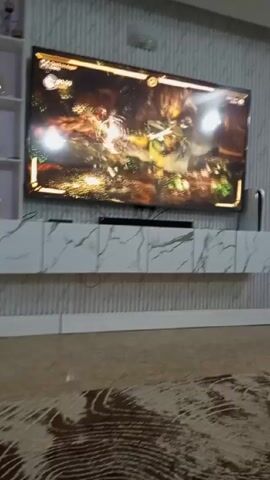 The video shows a person playing a fighting game on a large TV. The TV is mounted on a wall with vertical ribbed paneling above a low, white cabinet with a marble pattern. A PlayStation 5 is visible next to the TV. The game on the screen shows two characters fighting in a forest-like setting.  Sound effects from the game can be heard.