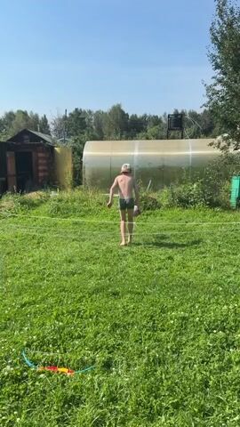 A young boy wearing green swim trunks and a baseball cap is playing with a soccer ball in a grassy yard. He is barefoot and appears to be practicing juggling the ball. Behind him is a large, transparent greenhouse and some trees. The sun is shining, creating a bright and summery atmosphere.