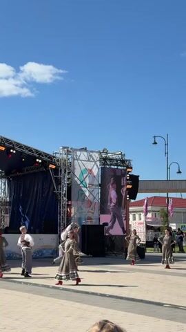A group of performers dressed in traditional clothing are dancing on a paved outdoor plaza in front of a stage. The stage has a large screen displaying a video of a person dancing. The sky is blue with a few white clouds. There are spectators in the foreground.