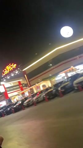 The video captures a nighttime scene outside a brightly lit building with Arabic signage that reads "Kabdah Al-Dahhan." A row of parked cars, mostly sedans and SUVs, is visible in the foreground, with orange traffic cones marking spaces. A black sedan turns into the frame from the left, its taillights illuminated. Two individuals are walking in the middle ground toward the building, which appears to be a restaurant or commercial establishment, with storefronts and seating areas visible. The building is adorned with red and white accents and illuminated with strings of lights and neon signs. A large, bright moon is visible in the upper right corner of the sky. The overall environment suggests a lively commercial area at night.