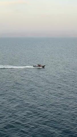 A small boat is moving from left to right across the frame, creating a white wake behind it. The boat is dark with a lighter colored cabin or top. The water is choppy and a deep blue. In the distance, a hazy, pale horizon separates the sea from a light colored sky. The sky is a soft gradient of pink and pale blue, suggesting it is either sunrise or sunset. There are no people visible in the video.