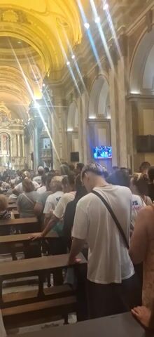 The video shows the interior of a large, ornate church filled with people. The church has high, arched ceilings and detailed architectural elements. Sunlight streams in through the windows. Most of the people are sitting in pews facing the altar, which is intricately decorated and brightly lit. There is a screen visible on one of the walls, showing what appears to be a live broadcast. People are wearing a variety of casual clothes, and some are wearing white shirts with logos. Some people are standing near the back of the church.