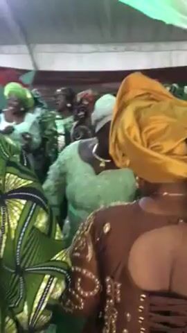 The video depicts a group of women dancing at an indoor event. The women are dressed in colorful and patterned clothing, with many wearing traditional headwraps in green, yellow, and other colors. The setting appears to be under a tent or a covered area, decorated with fabrics and floral arrangements. The women are moving and dancing together, creating a lively and celebratory atmosphere.