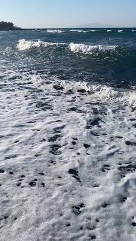 The video shows a close-up view of waves crashing onto a dark sand beach. The water is a deep blue and the waves are white and foamy. The camera is low to the ground, giving the impression of being at sea level. There are no people or objects visible in the video, only the natural environment of the beach and the ocean. The lighting suggests it is daytime, possibly late afternoon given the angle of the sun. In the background, there are hints of buildings on a hillside overlooking the water.