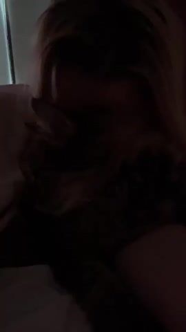 A person with blonde hair is holding a cat close to their face in a dark room. The person is wearing a light-colored shirt. The cat is a tabby pattern with dark fur and light markings. The person appears to be petting or nuzzling the cat. The background shows a dark curtain or wall.