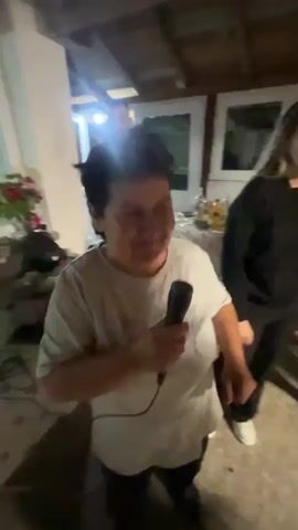 The video shows a small indoor gathering with several people in a room. A woman in a white t-shirt is holding a microphone and speaking or singing, seemingly enjoying herself. Behind her is a table laden with bottles and food, suggesting a party. Other individuals are moving around, chatting, and dancing. One woman with long hair has a purple flower clip in her hair. The room appears to be a garage or shed, with exposed wooden beams and windows in the background. The overall atmosphere seems festive and casual.