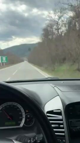 The video shows a view from the driver's seat of a car driving down a rural road. The road is asphalt, straight, and two lanes wide. Trees line the road on both sides, and a hill is visible in the distance. The sky is cloudy. A black sedan passes the car in the same direction. The car's dashboard is visible, showing the speedometer, fuel gauge, and other controls. Music is playing in the background.
