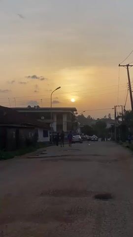 The video shows a street view at sunset. The sky is a mix of yellow and orange. In the distance, the sun appears as a bright white disk, partially obscured by clouds. The street is paved but cracked and potholed. On the left, there is a small building with a dark roof. A larger, multi-story building is behind it, equipped with satellite dishes. Several people are walking down the street, their figures silhouetted against the setting sun. Cars are parked on the side of the road. Power lines and street lights are visible above. A motorcyclist passes in the foreground.